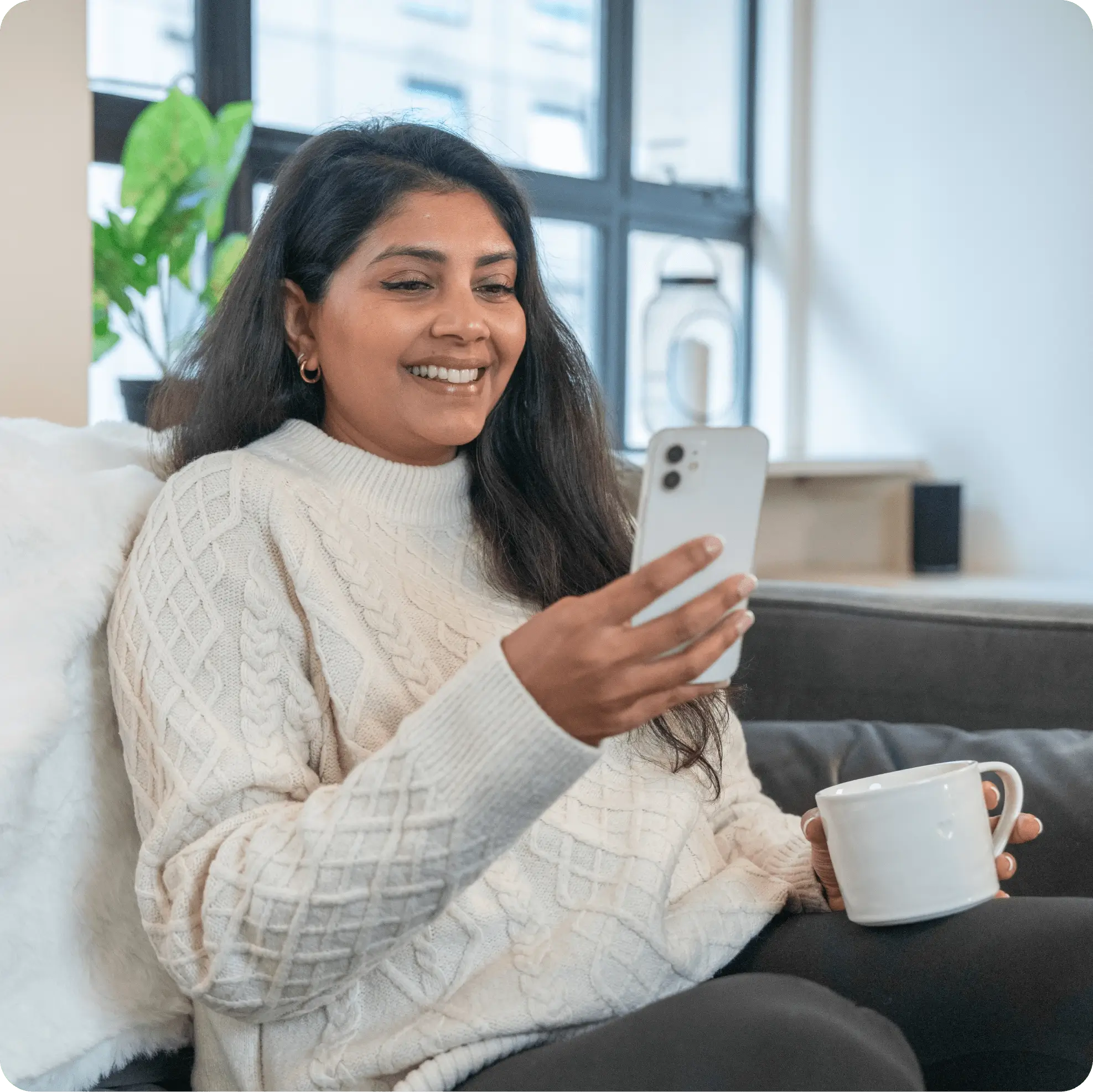 Smiling woman in a white sweater sitting on a couch holding a white mug and looking at her smartphone.