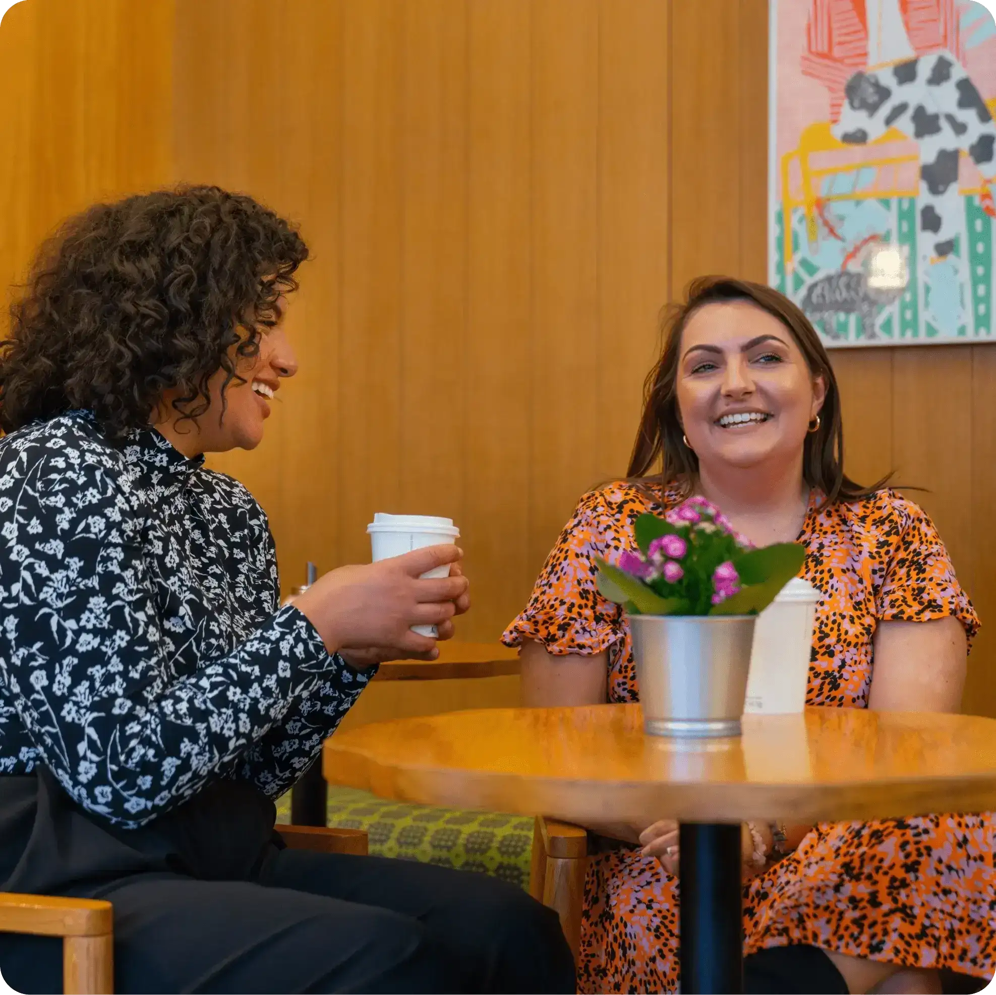Two women sitting at a round wooden table, smiling and chatting while holding white takeaway coffee cups, with a potted plant on the table.