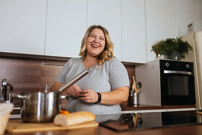 Smiling woman in gray shirt preparing food in a modern kitchen with a pot and bread on the counter.
