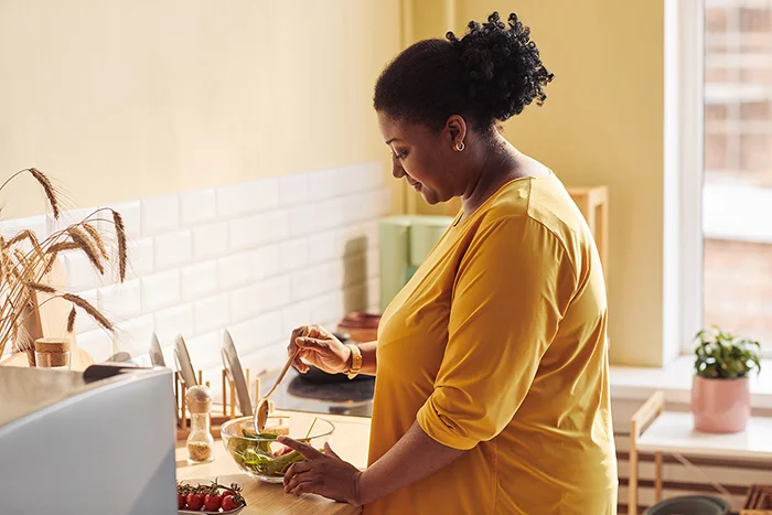 Woman in a yellow shirt stirring a salad in a bowl in a cozy kitchen with natural light.