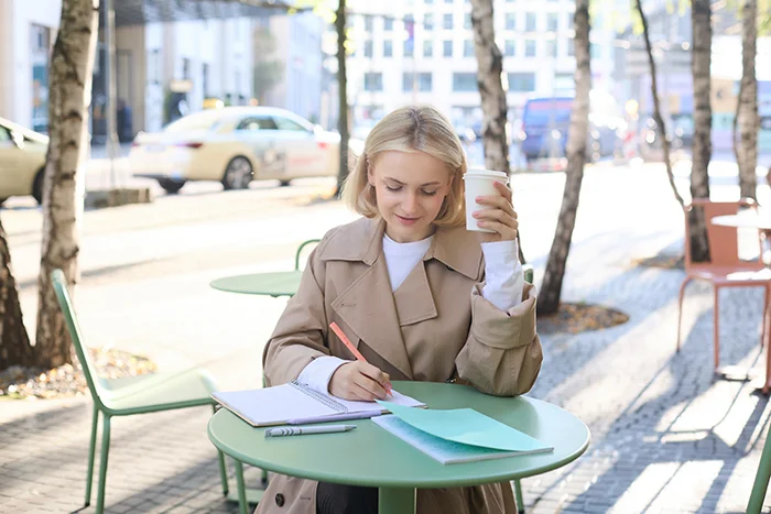 Young woman sitting at green outdoor table writing in a notebook while holding a coffee cup.