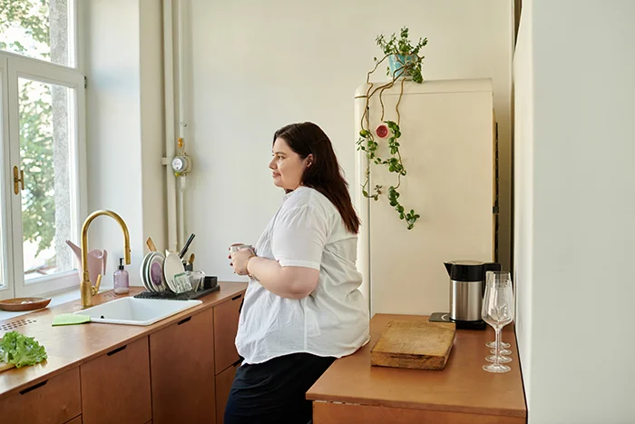 Plus-size woman in a white shirt sitting on a wooden kitchen counter holding a cup, looking out a window with natural light.
