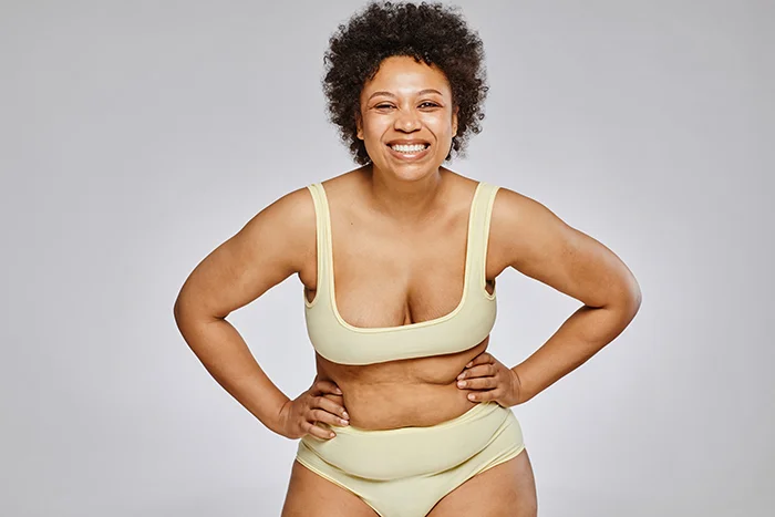 Smiling black woman with curly hair wearing a yellow bikini, standing with hands on hips against a light gray background.