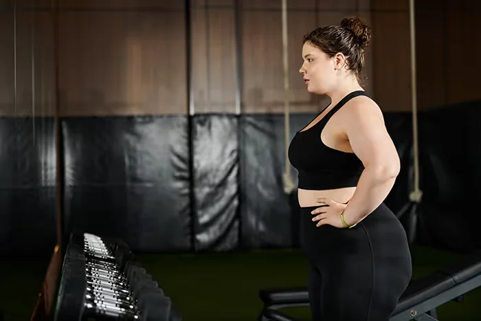 Young woman in black workout clothes standing with hands on hips, looking at a rack of dumbbells in a gym.