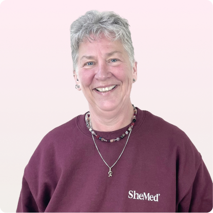 Smiling older woman with short gray hair wearing a maroon SheMed sweatshirt and beaded necklace against a light pink background.