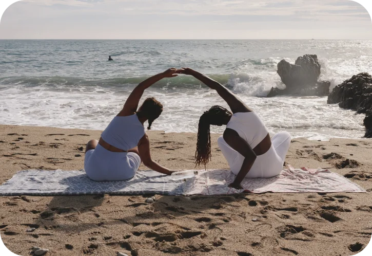 Two women in white workout clothes sitting on beach towels on the sand, stretching with arms raised and joined over their heads against a backdrop of ocean waves and rocks.