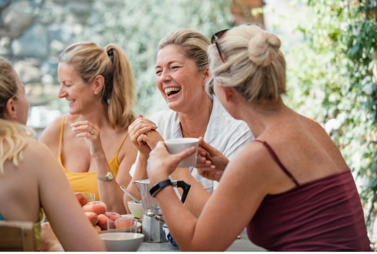 Four women sitting outdoors at a table, laughing and talking while enjoying coffee.