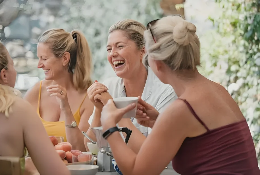 Four women laughing and enjoying coffee together at an outdoor table with fruit and a coffee pot.