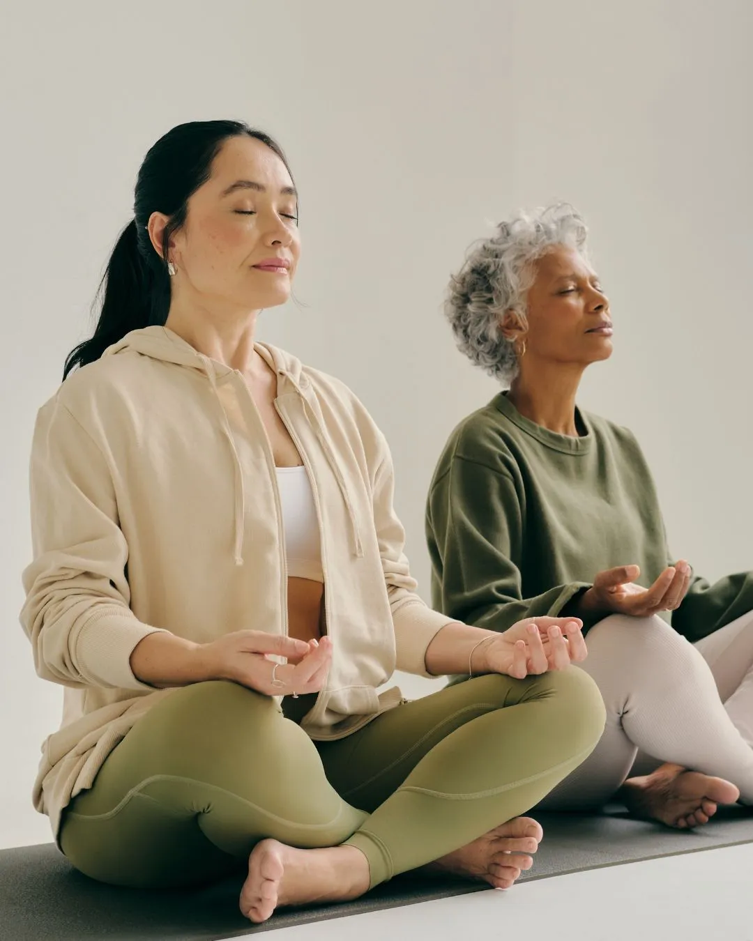 Two women sitting cross-legged on yoga mats with eyes closed, meditating indoors in relaxed posture.
