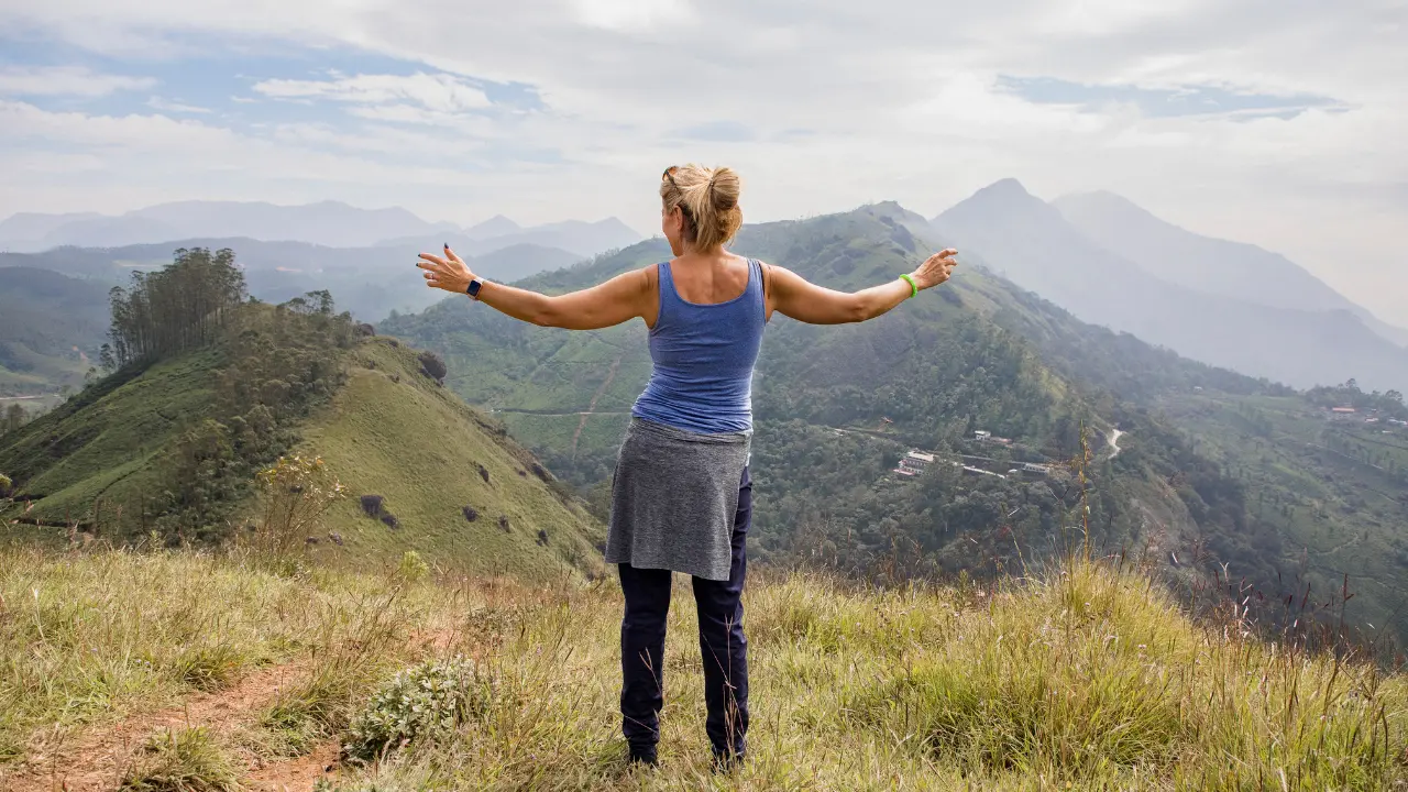 Woman with outstretched arms standing on a grassy hill overlooking misty mountains under a partly cloudy sky.