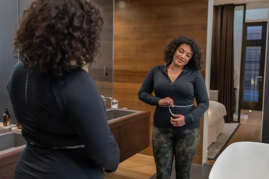 Woman measuring waist circumference in bathroom mirror using measuring tape for fitness tracking.