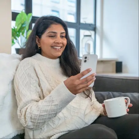 Woman relaxing on couch at home holding smartphone and coffee mug with plant in background.