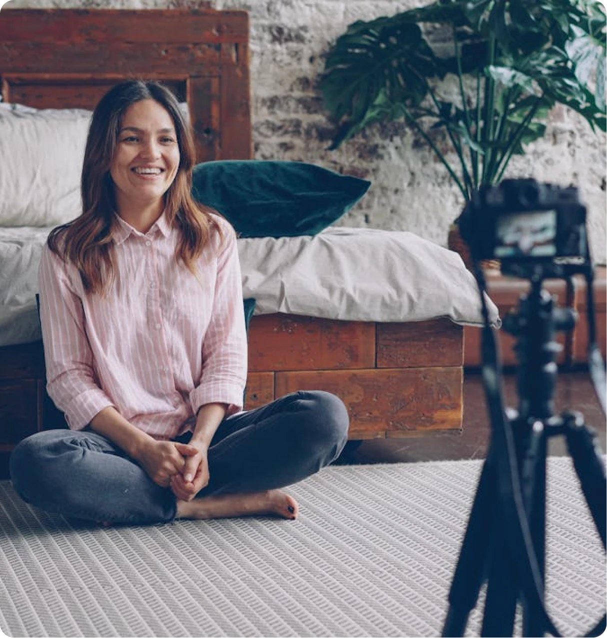 Smiling woman sitting cross-legged on a carpeted floor recording a video with a camera on a tripod in a cozy bedroom.