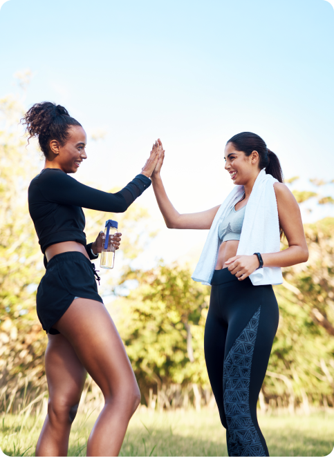 Two women in workout attire giving each other a high five outdoors, one holding a water bottle and the other with a towel around her neck.