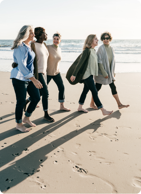 Five diverse women walking barefoot on a sunny beach near the ocean, smiling and enjoying each other's company.