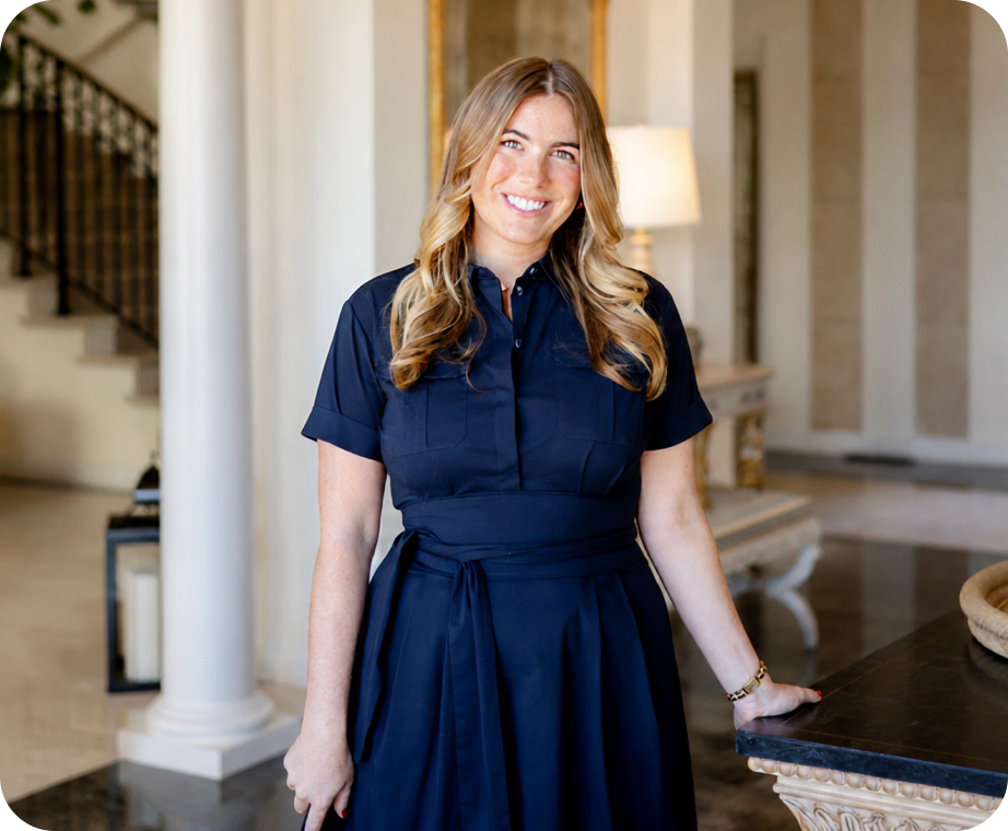 Smiling woman with long blonde hair wearing a navy dress, standing indoors by a table and column.