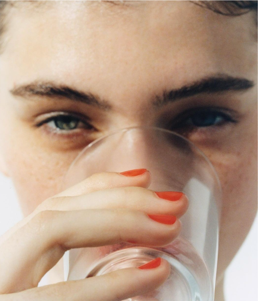 Close-up of a person with red nail polish holding a glass of water near their face.