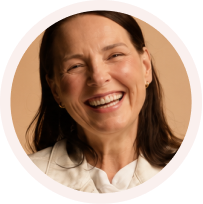 Smiling woman with long brown hair wearing a white top against a beige background.