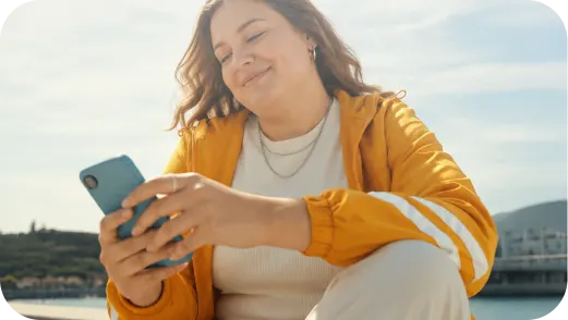 Smiling woman in yellow jacket using a smartphone outdoors by water and hills on a sunny day.