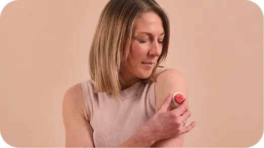 Woman applying an at-home blood test device to her upper arm against a beige background.