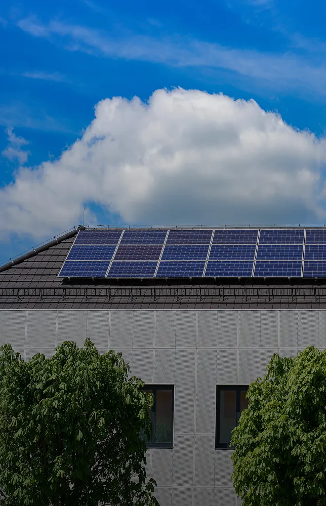 Solar panels installed on the roof of a modern building with two trees in front and a partly cloudy blue sky above.