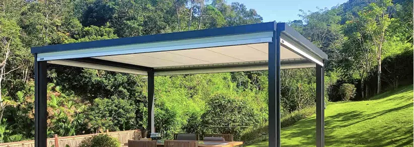 Outdoor patio pergola with retractable roof over a wooden table and chairs, set in a green garden with trees and grass.