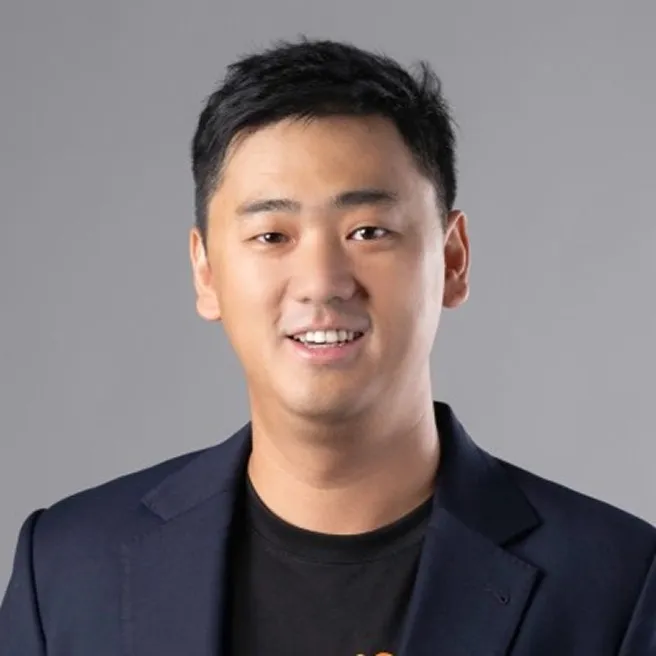 Portrait of a smiling young man with short black hair wearing a dark blazer and black shirt against a plain gray background.