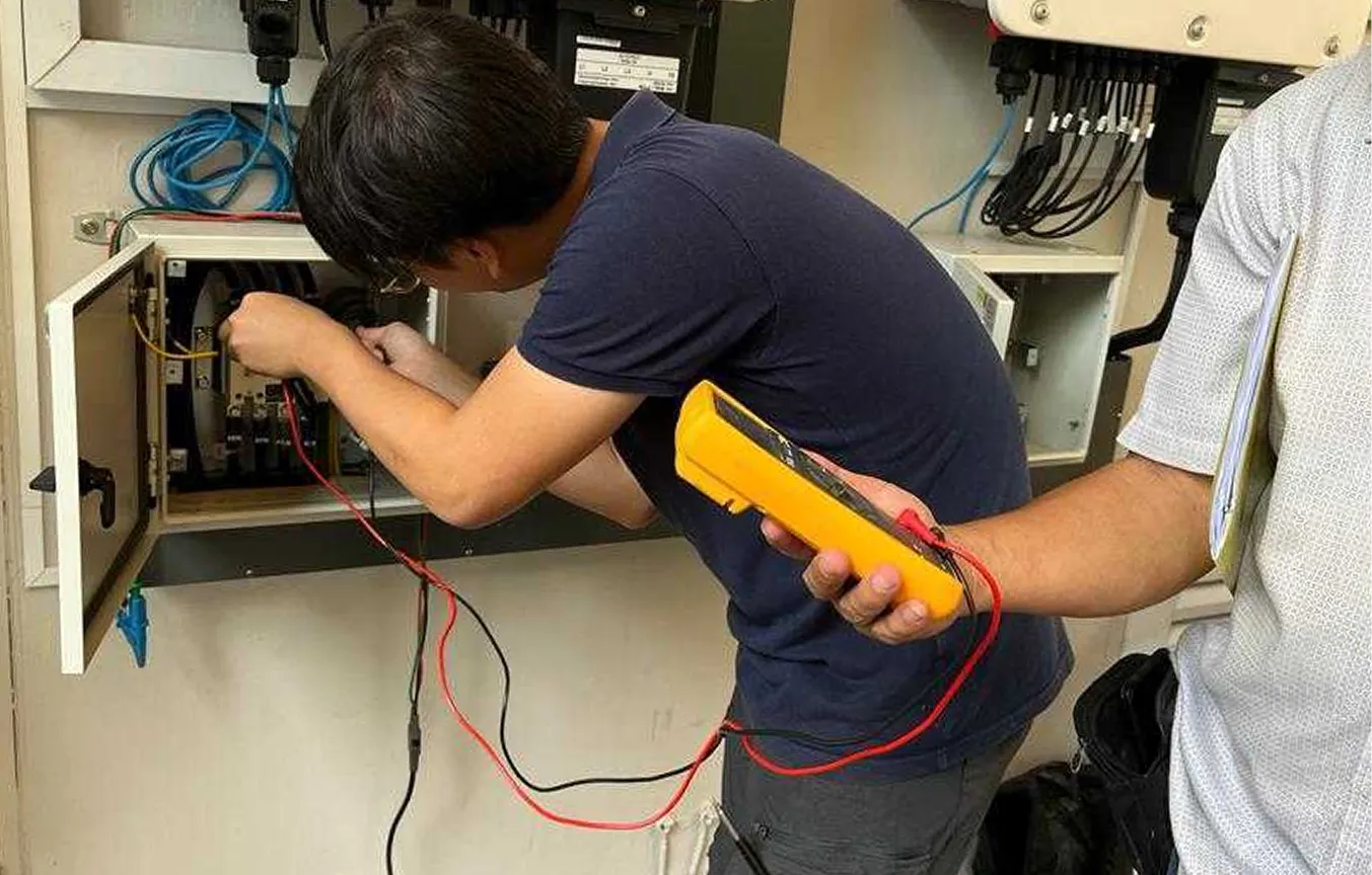 A technician in a blue shirt adjusting wiring inside an open electrical panel while another person holds a yellow multimeter.