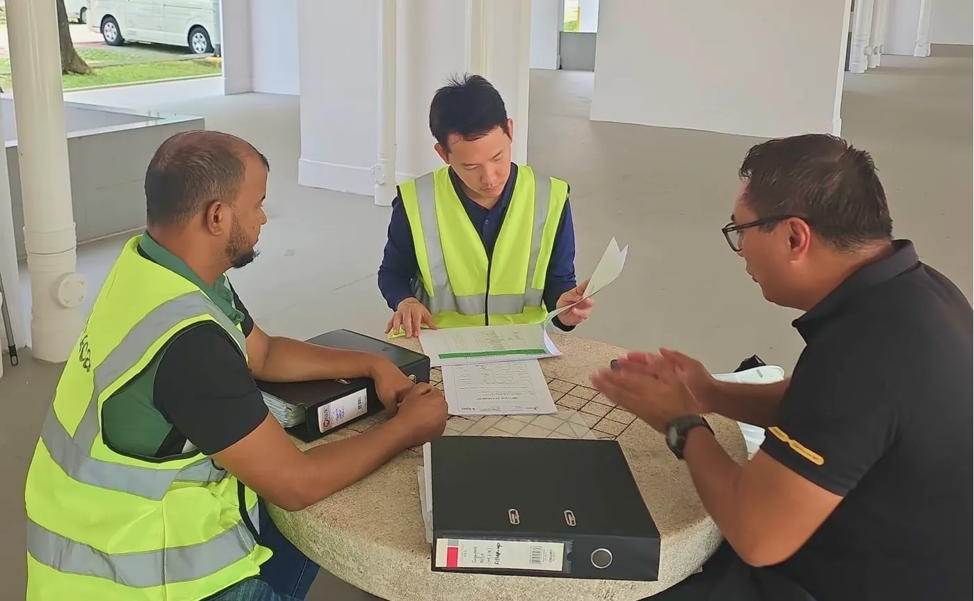 Three men at a round table reviewing documents, two wearing yellow safety vests and one in a black shirt.