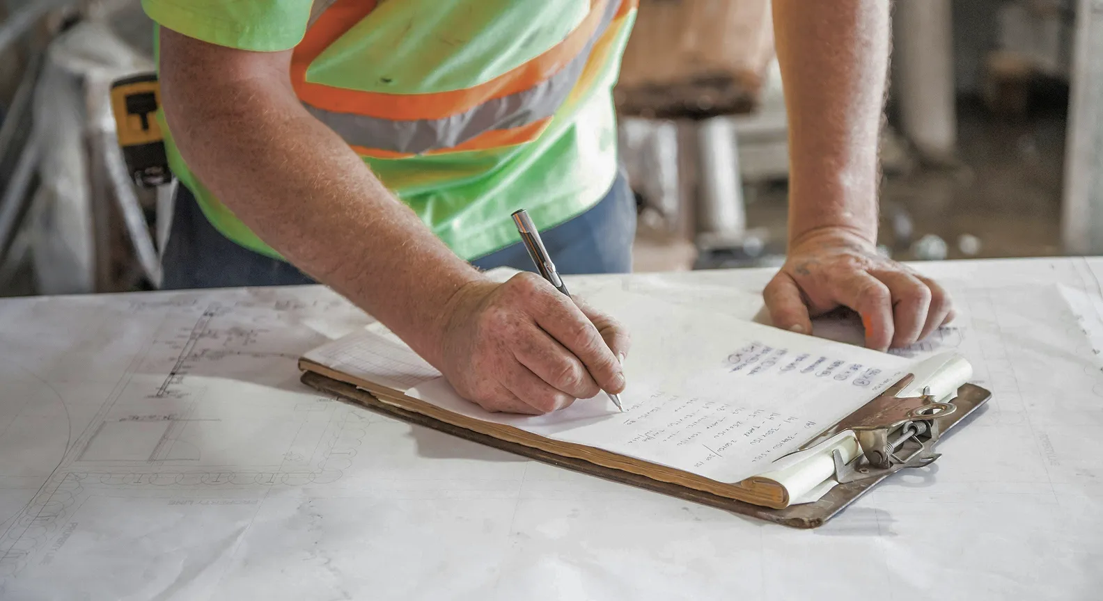Construction worker wearing a green safety vest writing on a clipboard with documents on a work table.