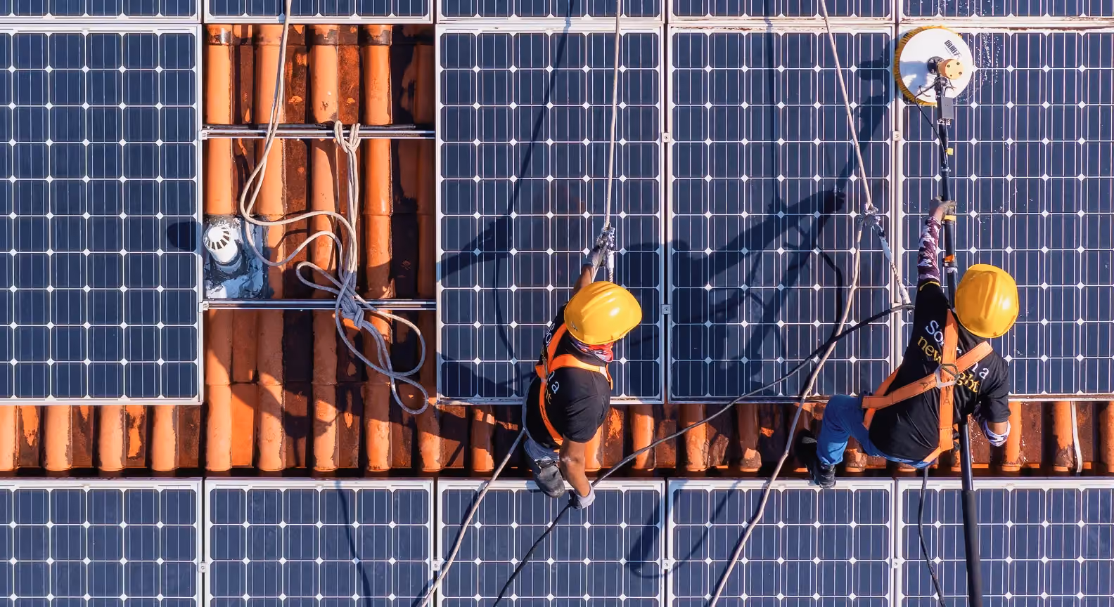 Two workers wearing safety harnesses and helmets cleaning solar panels installed on a tiled roof.