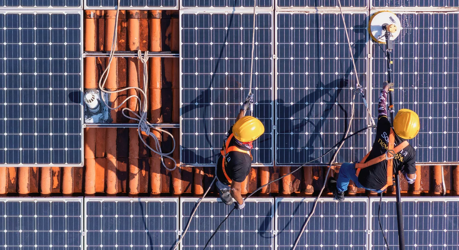 Two workers wearing safety harnesses and helmets cleaning solar panels installed on a tiled roof.