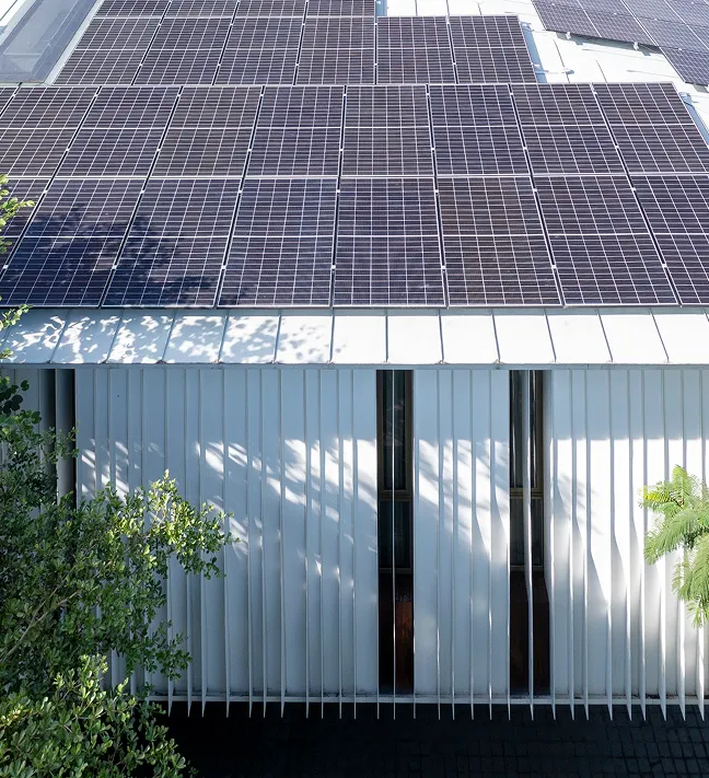 Rooftop covered with multiple solar panels above a white building facade with vertical slats and narrow windows, partially shaded by nearby trees.