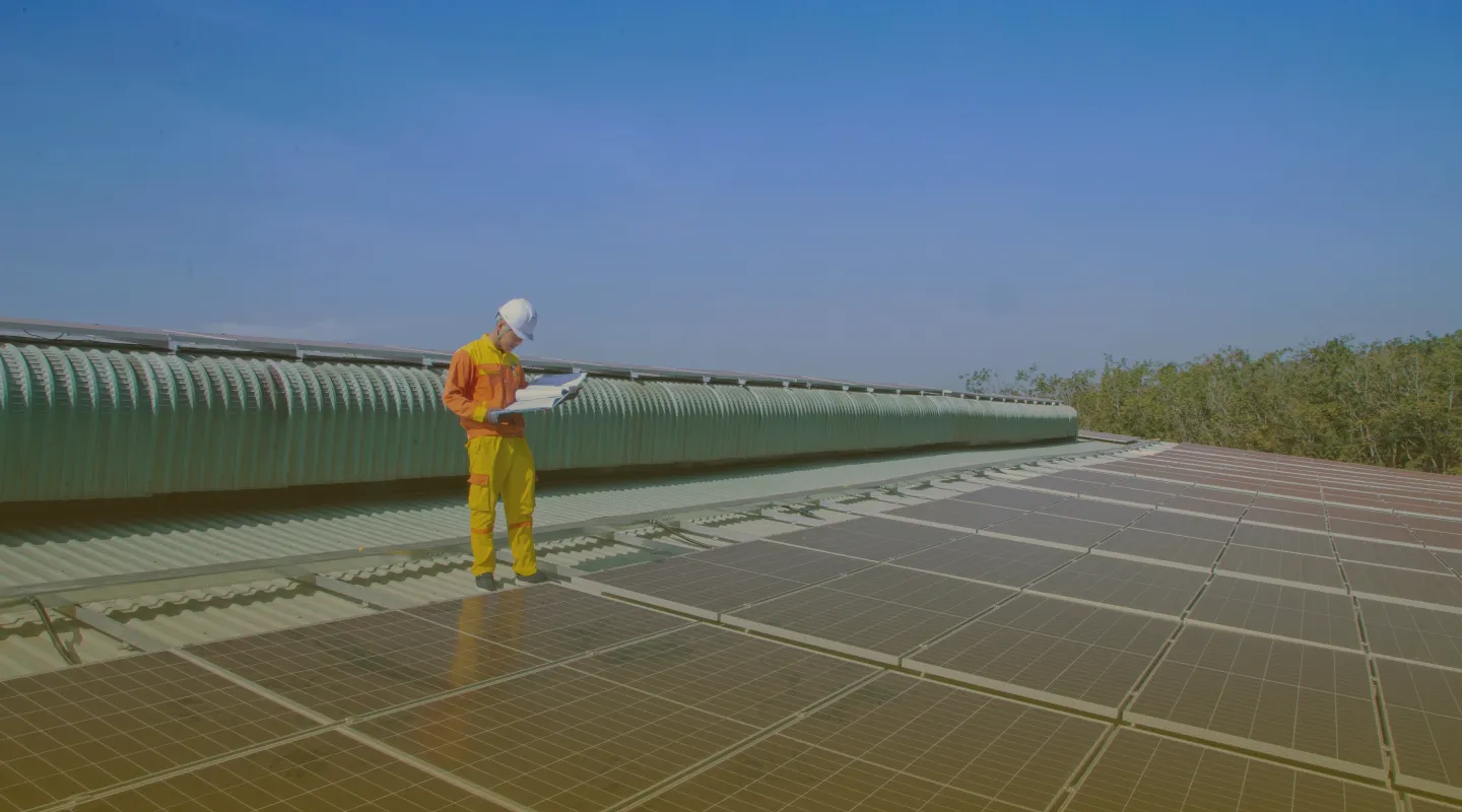 Worker in yellow and orange safety gear and helmet inspecting solar panels on a rooftop under a clear blue sky.