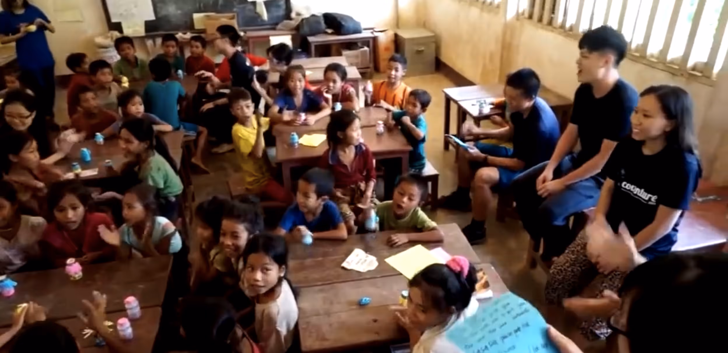 A lively classroom filled with children sitting at wooden desks and three young adults seated by the window, interacting and smiling.