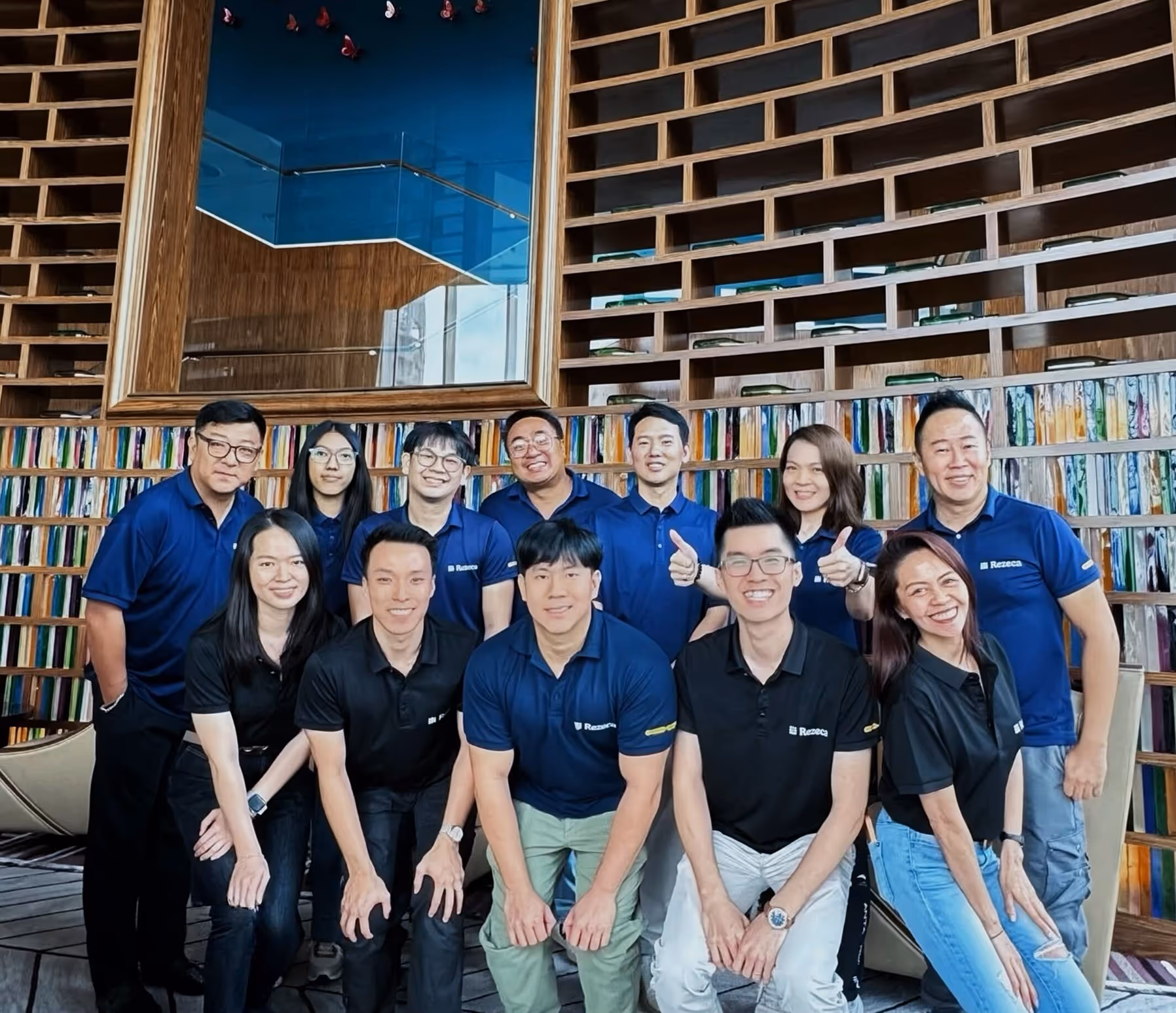 Group of twelve people wearing black and navy blue polo shirts posing indoors in front of a decorative wooden wall with colorful books or panels.
