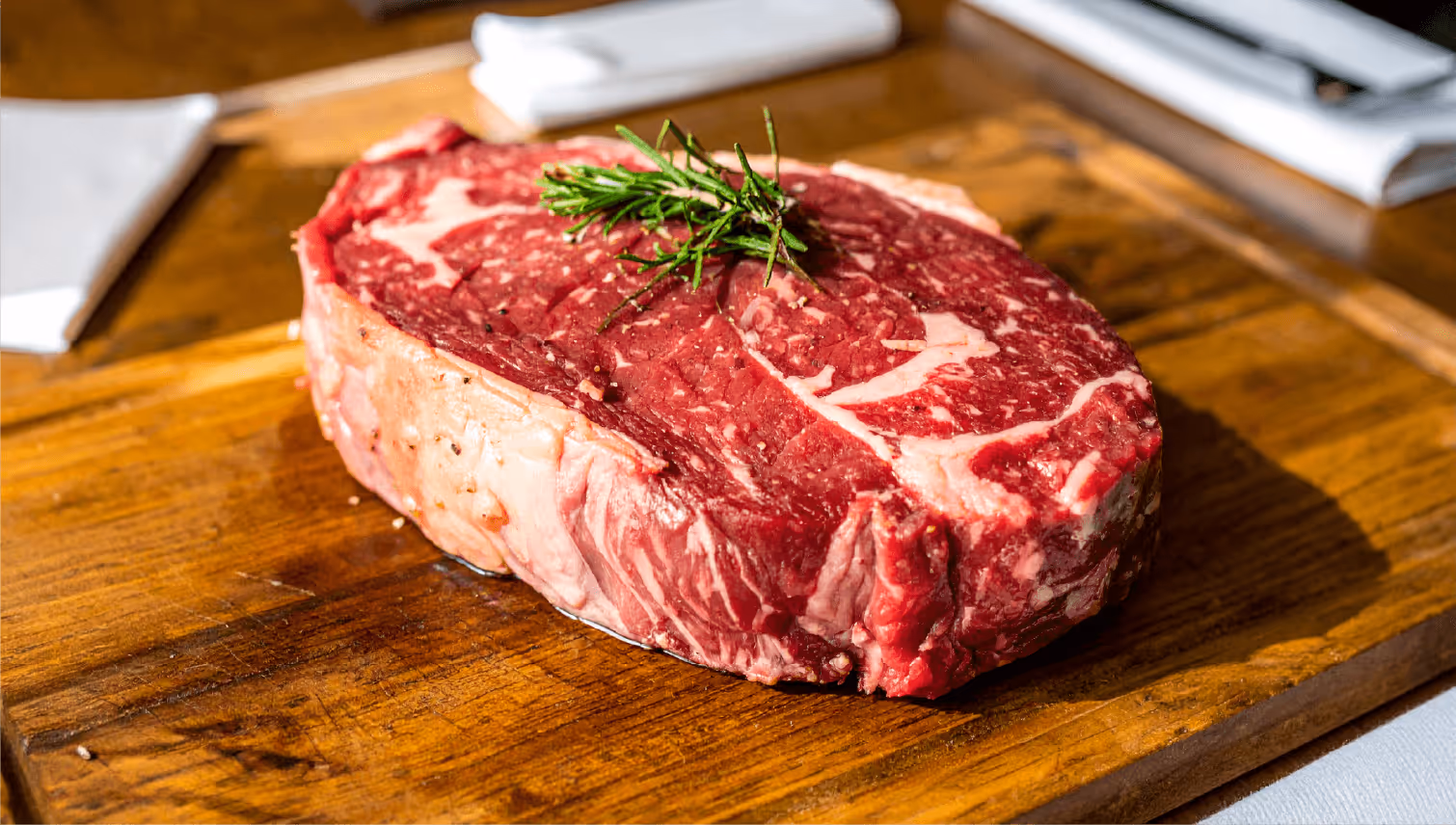 Raw ribeye steak with marbling and a sprig of rosemary on a wooden cutting board.