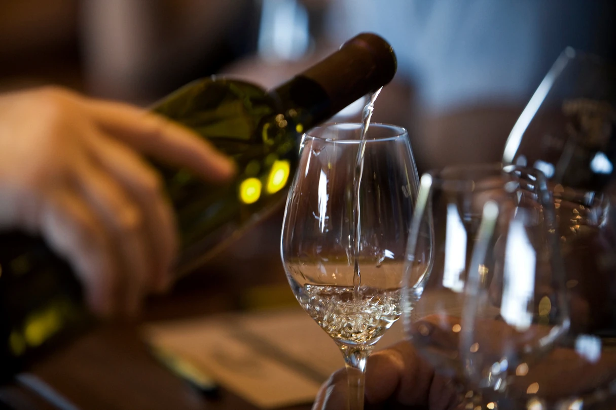 Close-up of a hand pouring white wine into a glass with other empty wine glasses blurred in the background.