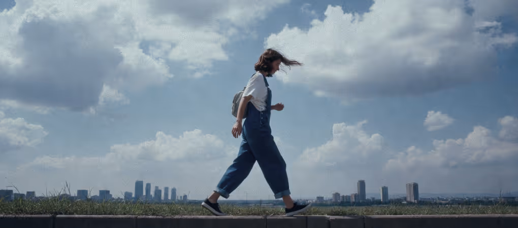 Person in blue overalls walking on a ledge with a city skyline and cloudy sky in the background.