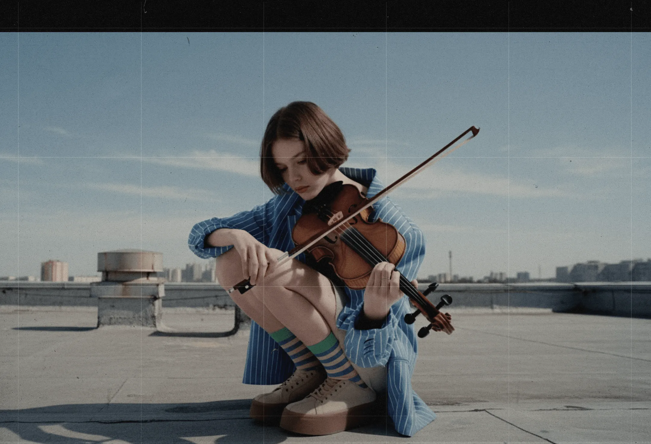 Young woman in a blue striped outfit playing violin while squatting on a rooftop under a clear sky.