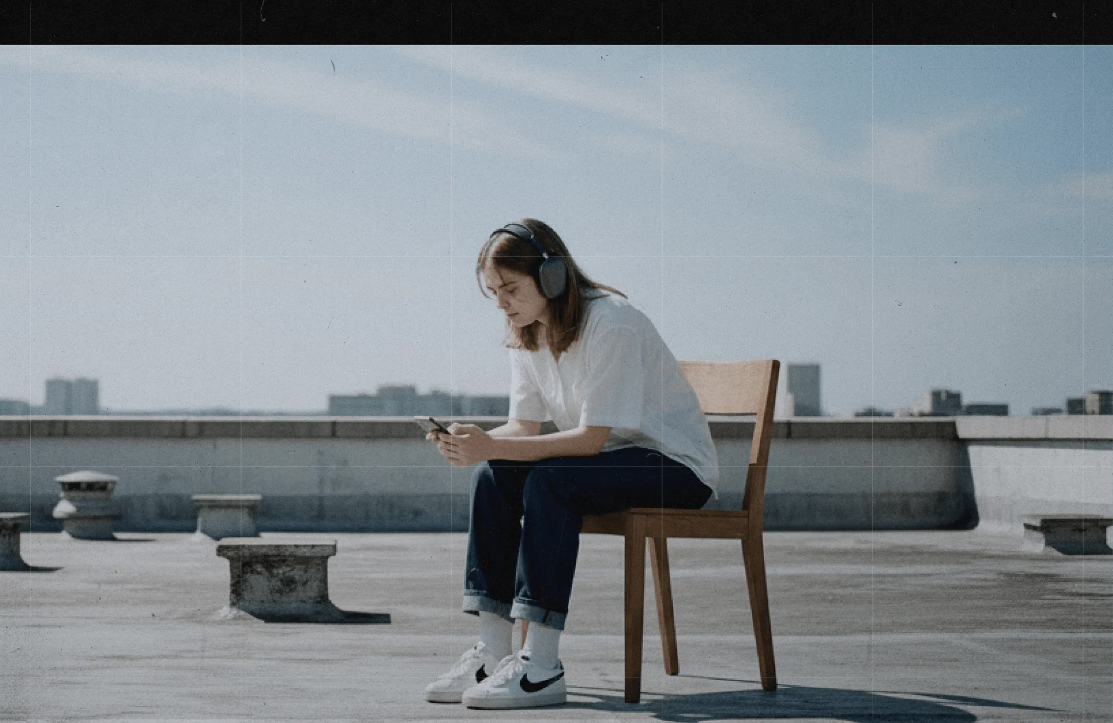 Person wearing headphones and casual clothes sitting on a wooden chair on a rooftop looking at a smartphone.