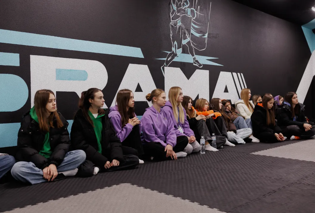 Group of young women sitting cross-legged on mats inside a gym, against a wall with large 'DRAMA' text and a boxing figure graphic.