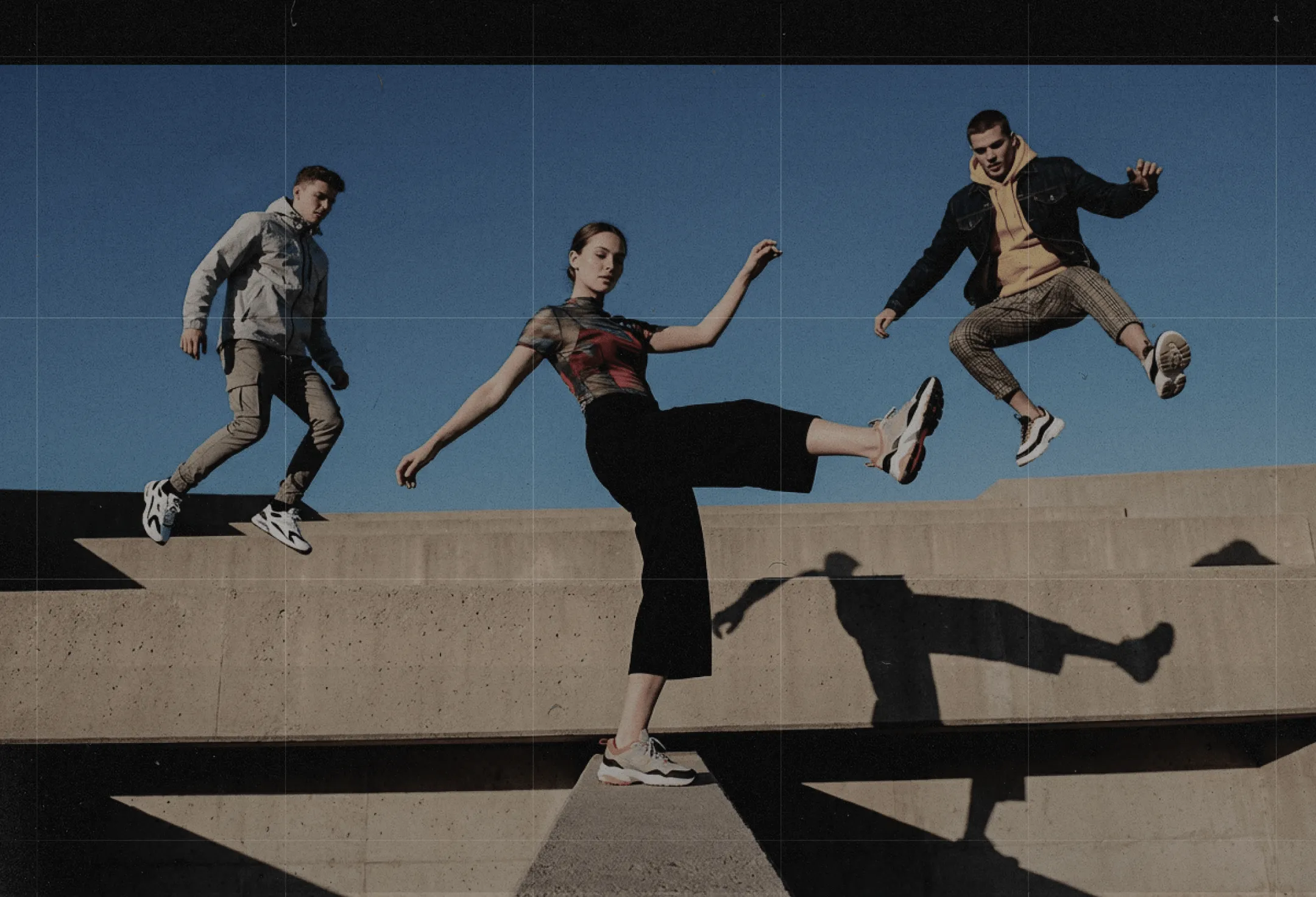 Three young people jumping and balancing on concrete structures under clear blue sky.