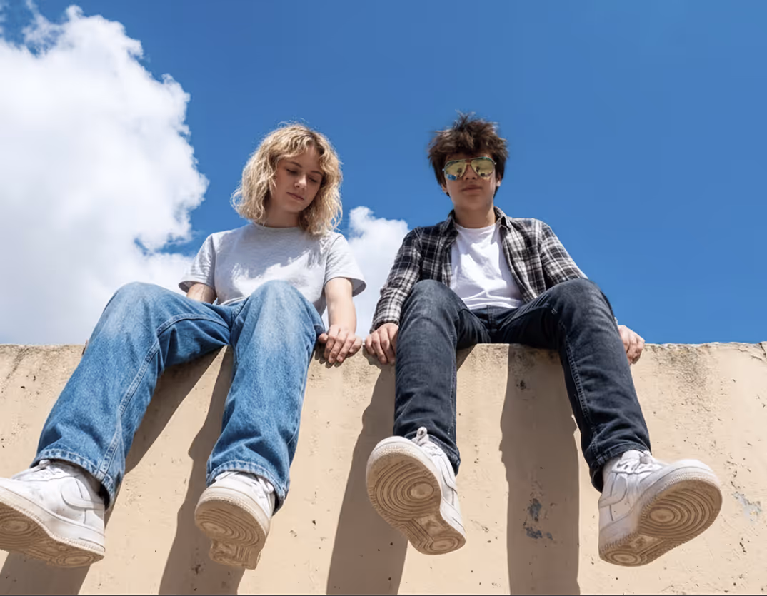 Two young people sitting on a ledge against a blue sky, holding hands, one wearing sunglasses.
