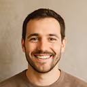 Smiling man with short dark hair and beard wearing a brown shirt against a beige background.