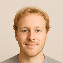 Portrait of a young man with short blond hair and light facial hair, smiling slightly against a plain beige background.