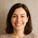 Smiling woman with shoulder-length brown hair wearing a white shirt against a beige background.