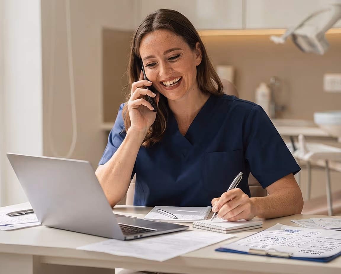 Smiling woman in navy scrubs talking on phone while writing notes at a desk with laptop and documents.