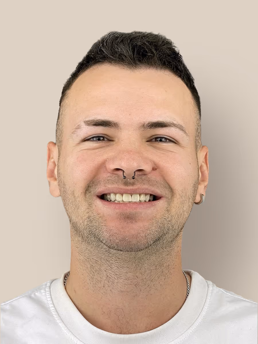 Smiling man with a septum nose piercing and short dark hair wearing a white shirt against a beige background.