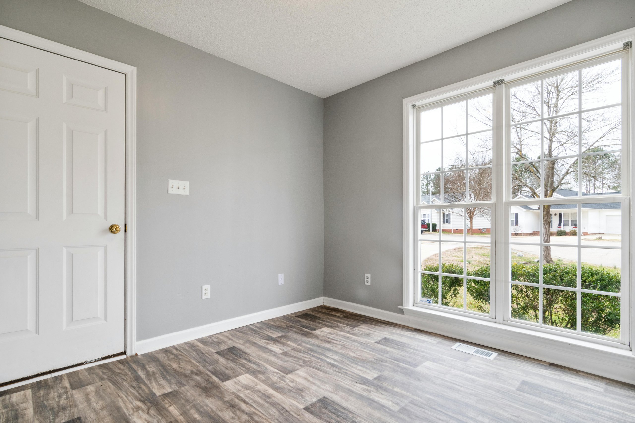 Empty room with gray walls, wood-patterned flooring, a white door with brass knob, and a large window looking out onto a suburban street.